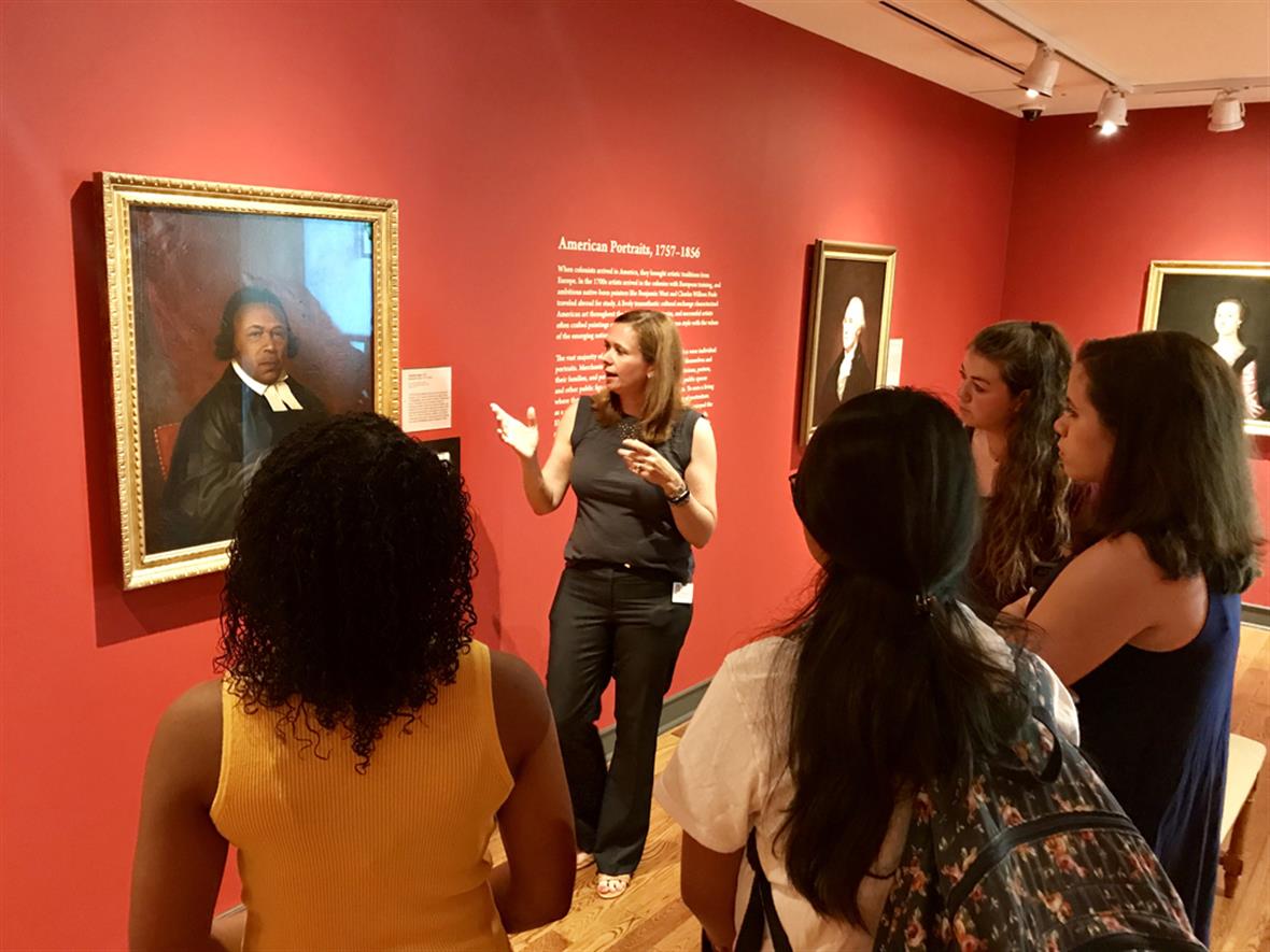 Heather Campbell Coyle stands in front of a painting in the museum while addressing a group of students.