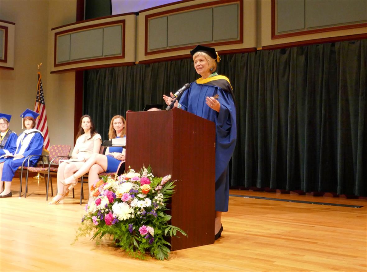 Debra Hess Norris standing behind a podium on a stage talking to an audience and wearing a convocation robe and hat.