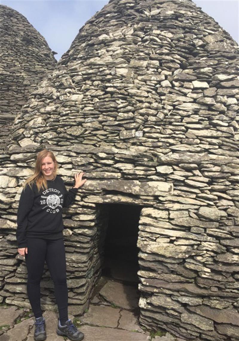Caitlin Hutchison standing in front of a beehive-shaped hut made of rocks