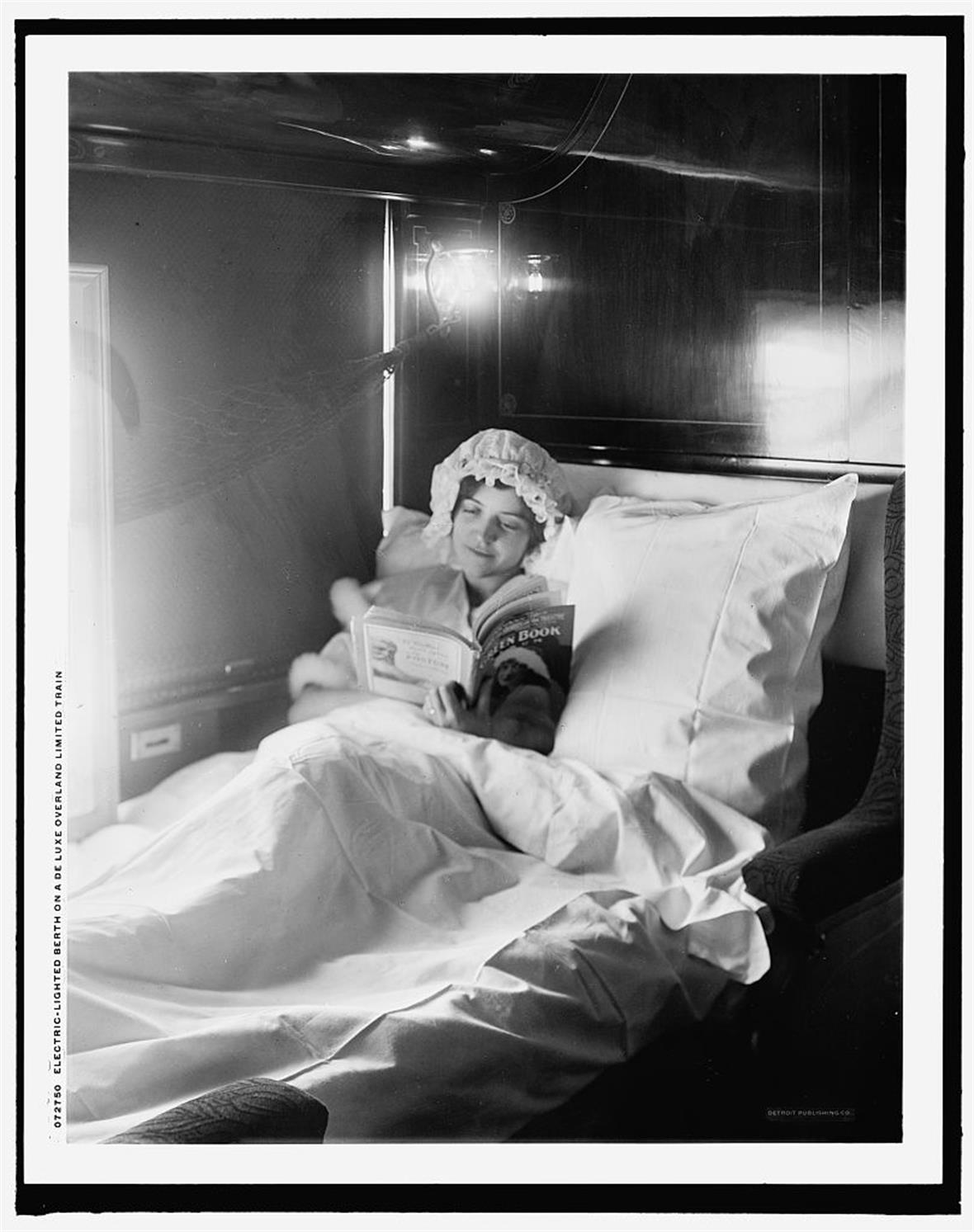 a black and white photograph of a woman reading a book in a bed on a train. There is a light behind her, illuminating the room.