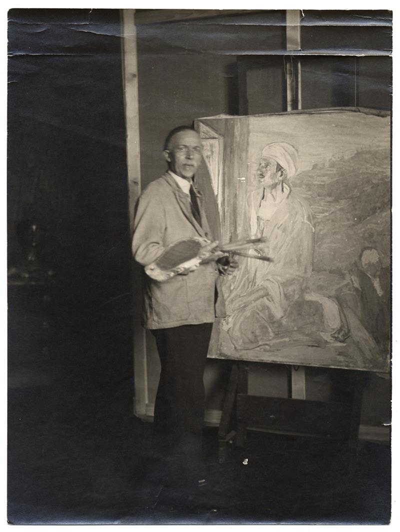A man holding an easel and paintbrushes standing next to a canvas