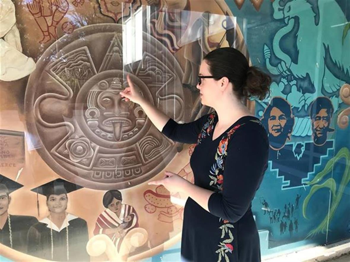 Rachel Zimmerman points to a circular wall relief hanging behind a glass case that appears to be of Native or indigenous origin.